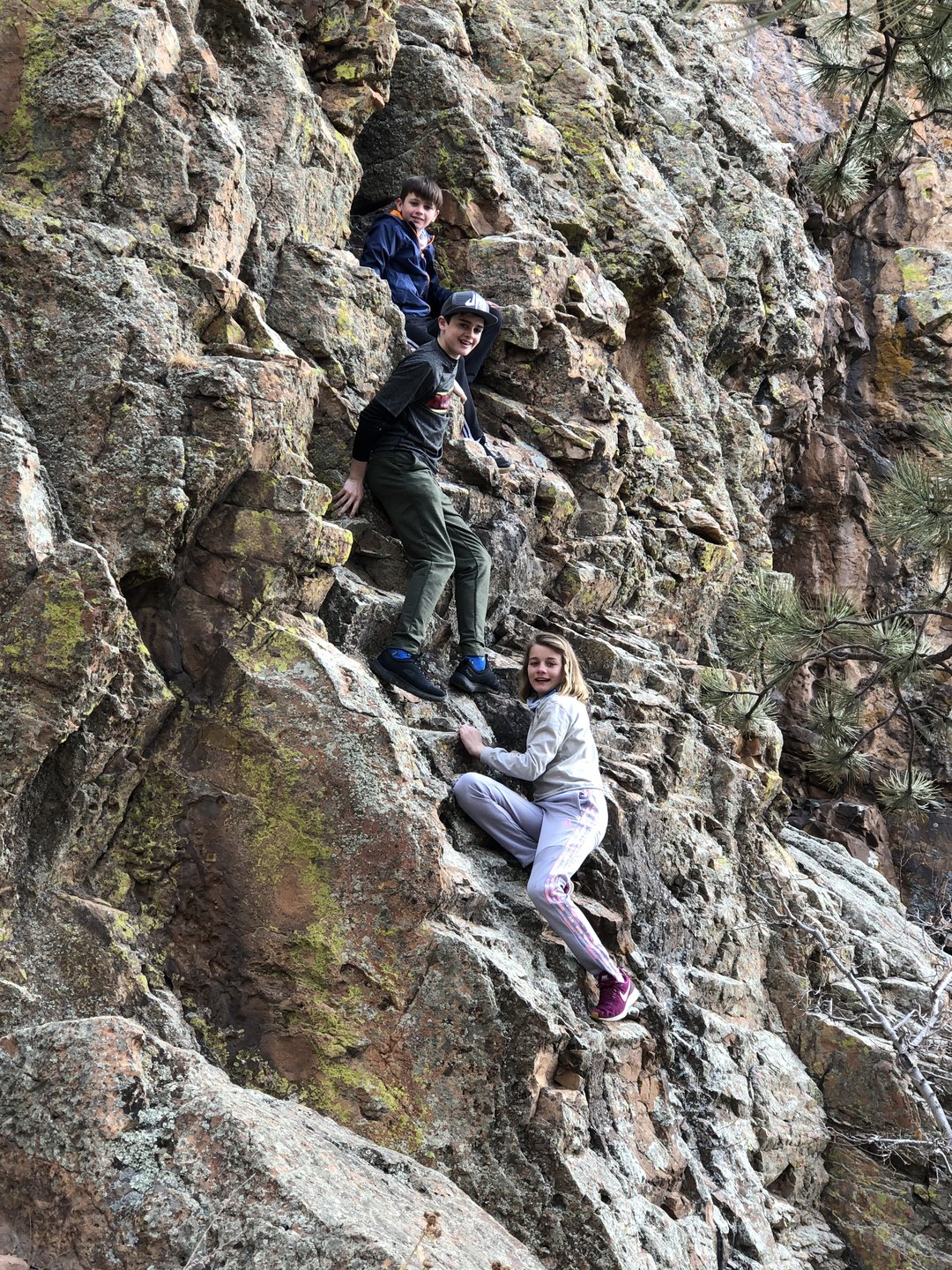 Three kids scrambling up a lichen-covered outdoor rock face